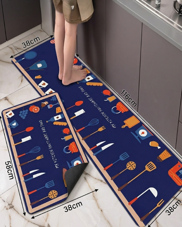A kitchen floor mat set shown in a real kitchen environment, with a person's legs visible stepping onto the mat. The set includes a long runner mat and a smaller square mat, both dark blue with colorful kitchen utensil illustrations and white text.