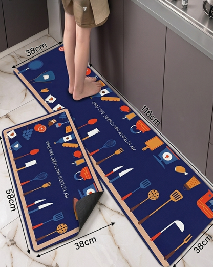 A kitchen floor mat set shown in a real kitchen environment, with a person's legs visible stepping onto the mat. The set includes a long runner mat and a smaller square mat, both dark blue with colorful kitchen utensil illustrations and white text.